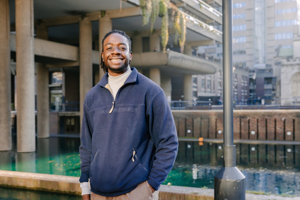 picture of a tall black man with locs and a beard, stood in front of the waters and buildings at the barbican centre